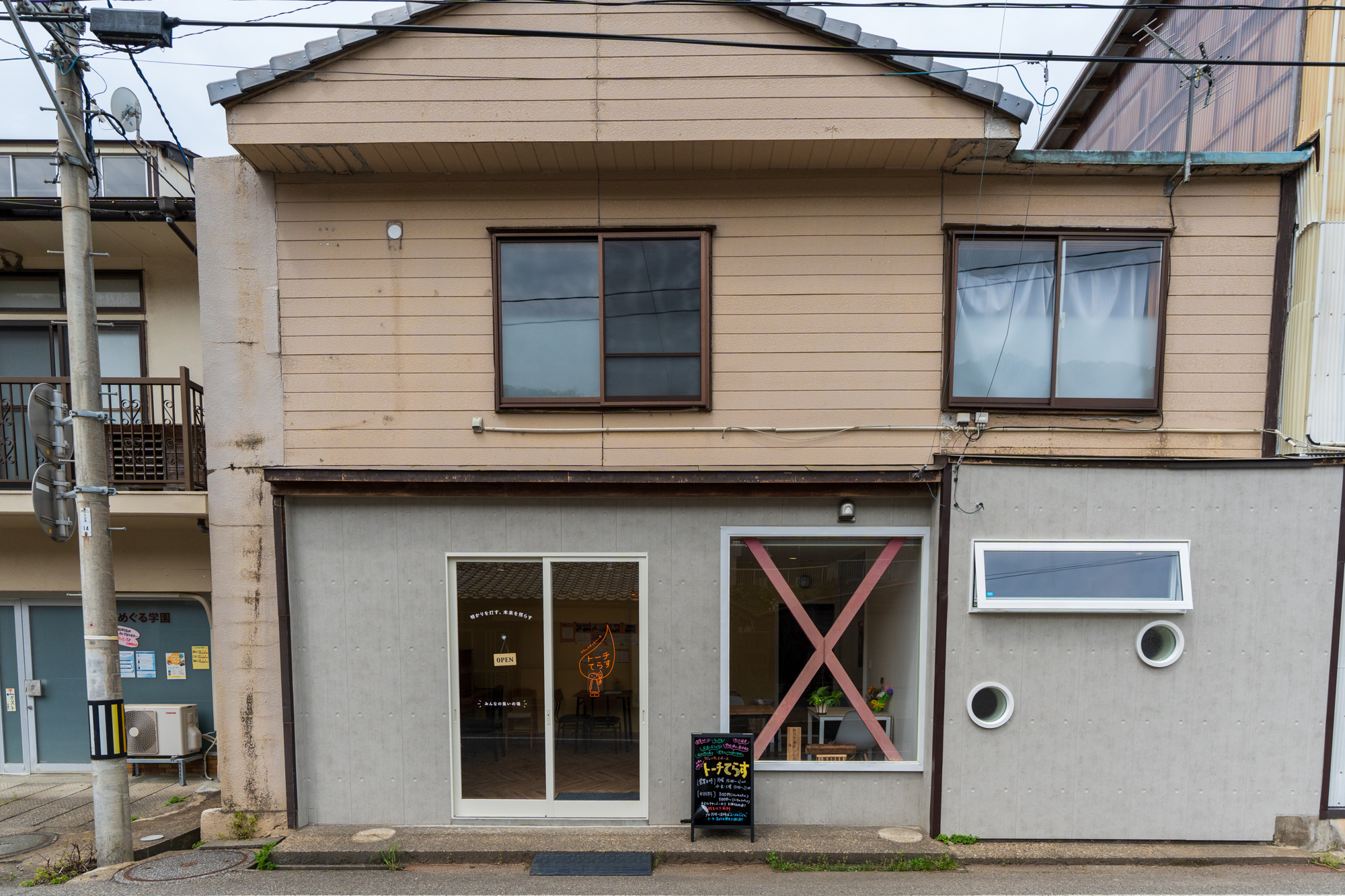 トーチてらすの建物外観。2階は木目のサイディング、1階はグレー系の外壁とガラス入口、看板が置かれた歩道