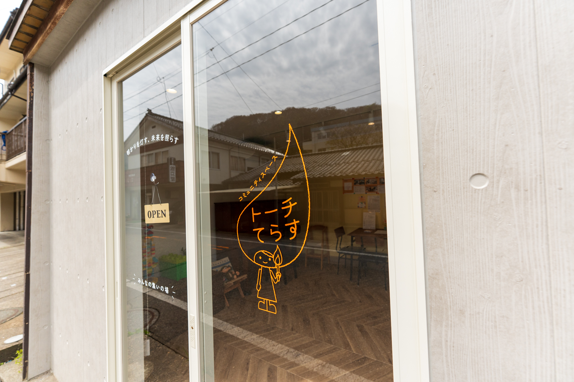 エントランスのガラス扉。炎のロゴと「コミュニティスペース トーチてらす」「明かりを灯す、未来を照らす」の文字、店内のヘリンボーン床がうかがえる